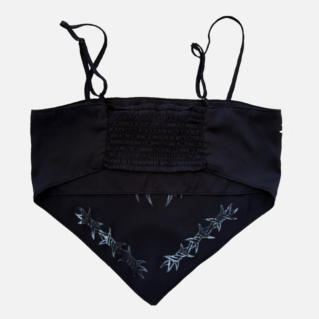 A black triangle bandana-style top with thin shoulder straps, a smocked chest panel, and silver barbed wire print along the lower front, shown against a light gray background.
