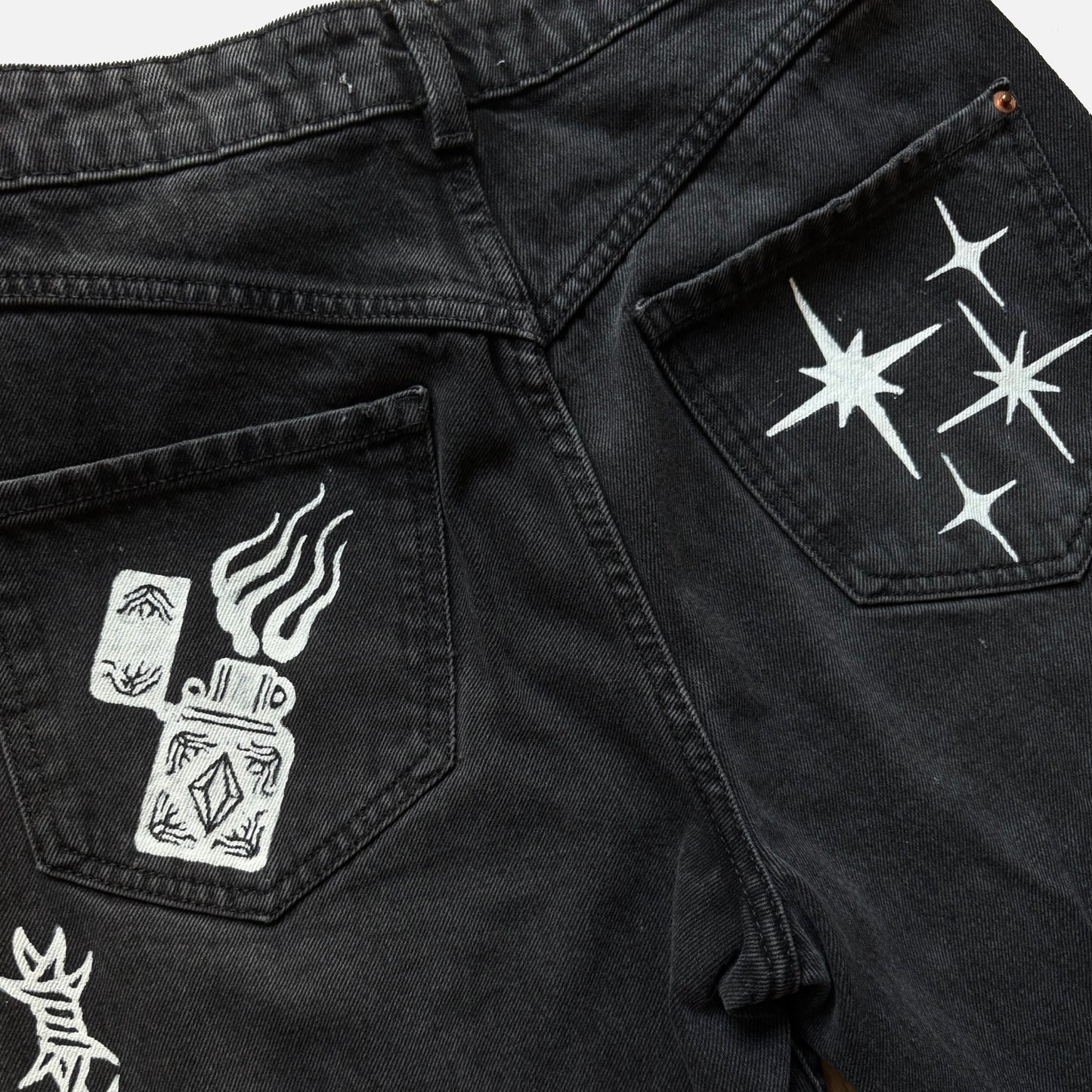 Close-up of black denim jeans showing the back pockets with white printed graphics, including a zippo lighter design on one pocket and starbursts on the other.