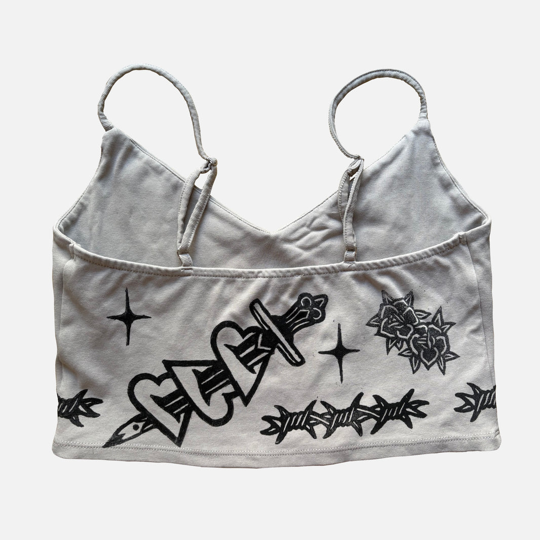 A beige cropped camisole top with black graphic prints of a dagger, roses, barbed wire, and stars, displayed against a light gray background.