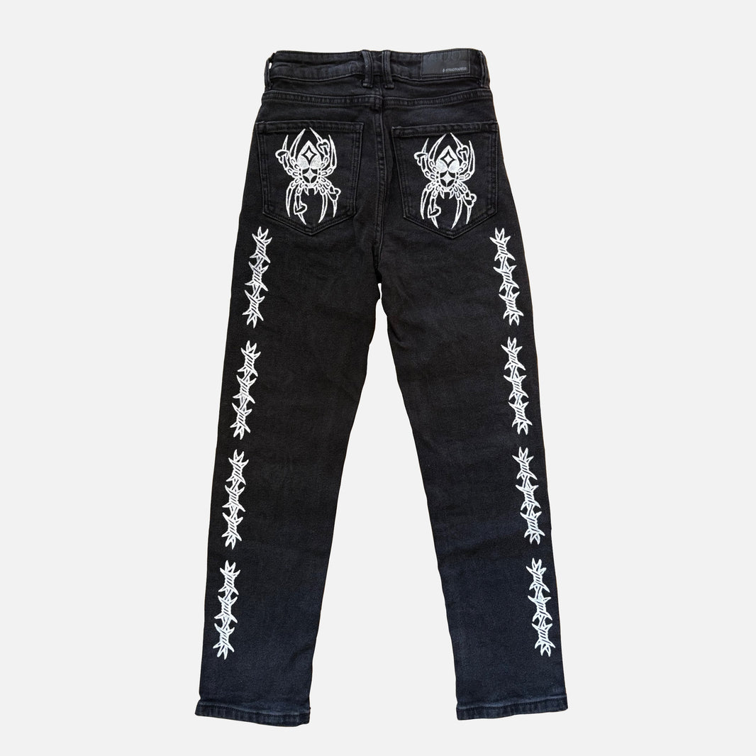 Black denim jeans featuring white printed gothic spider designs on the back pockets and vertical white printed barbed wire patterns down both legs, shown against a light gray background.