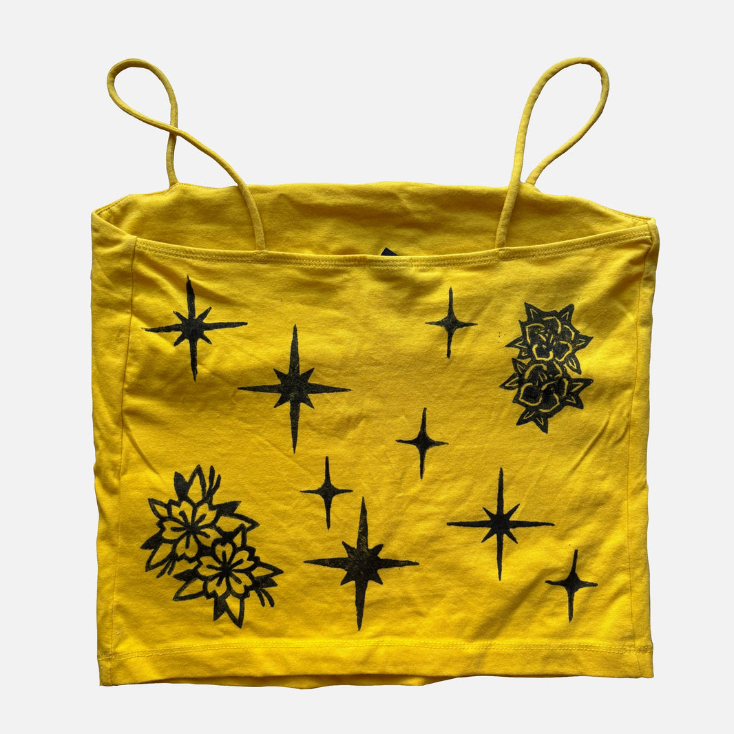 A yellow crop top with thin shoulder straps, featuring black starburst and floral line-art patterns printed across the front, shown against a plain light gray background.