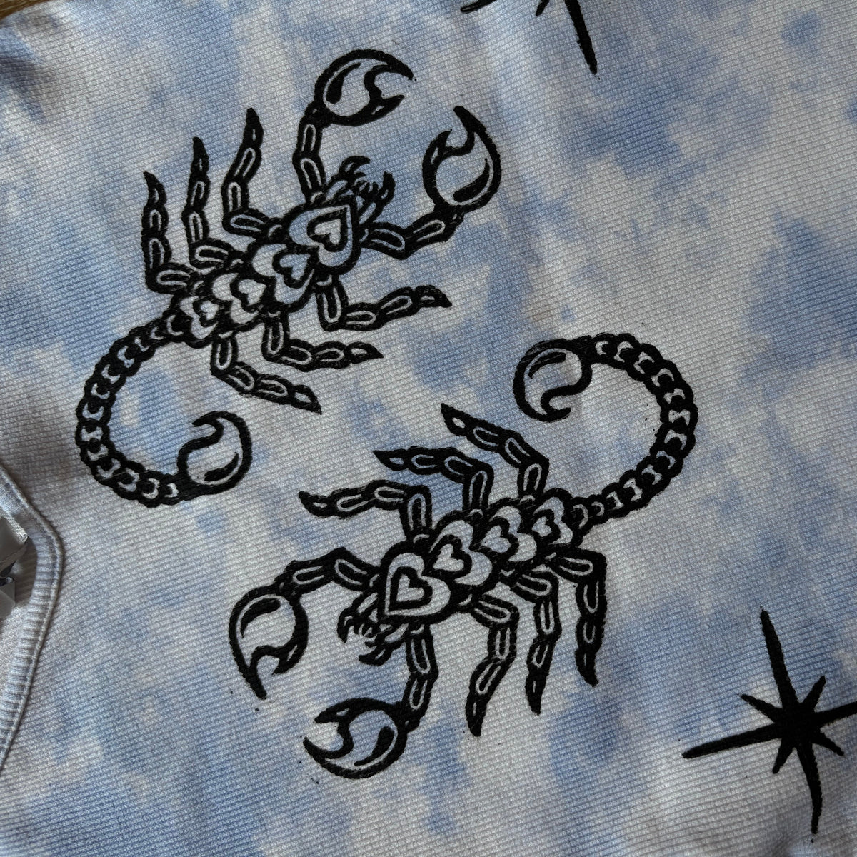 A close-up of a light blue and white tie-dye fabric featuring black line-art illustrations of scorpions and starbursts