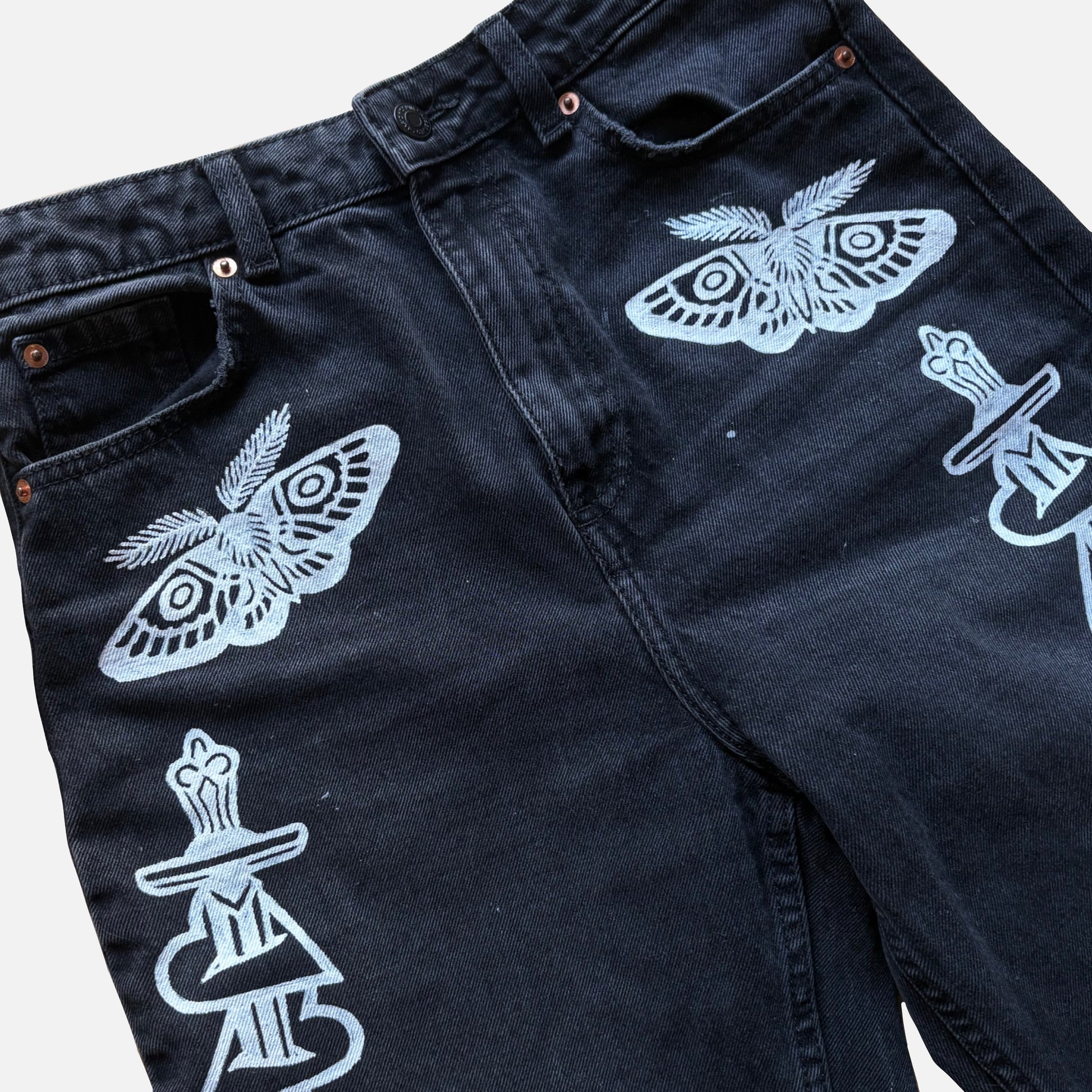 Close-up of dark washed denim jeans with white graphic prints of moths and dagger motifs on the front pockets, shown against a light gray background.