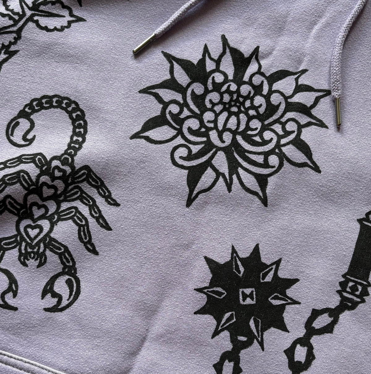 A close-up of a light purple hoodie featuring black graphic prints of a scorpion, a large flower, a flail, and a scorpion, with purple drawstrings visible at the top.