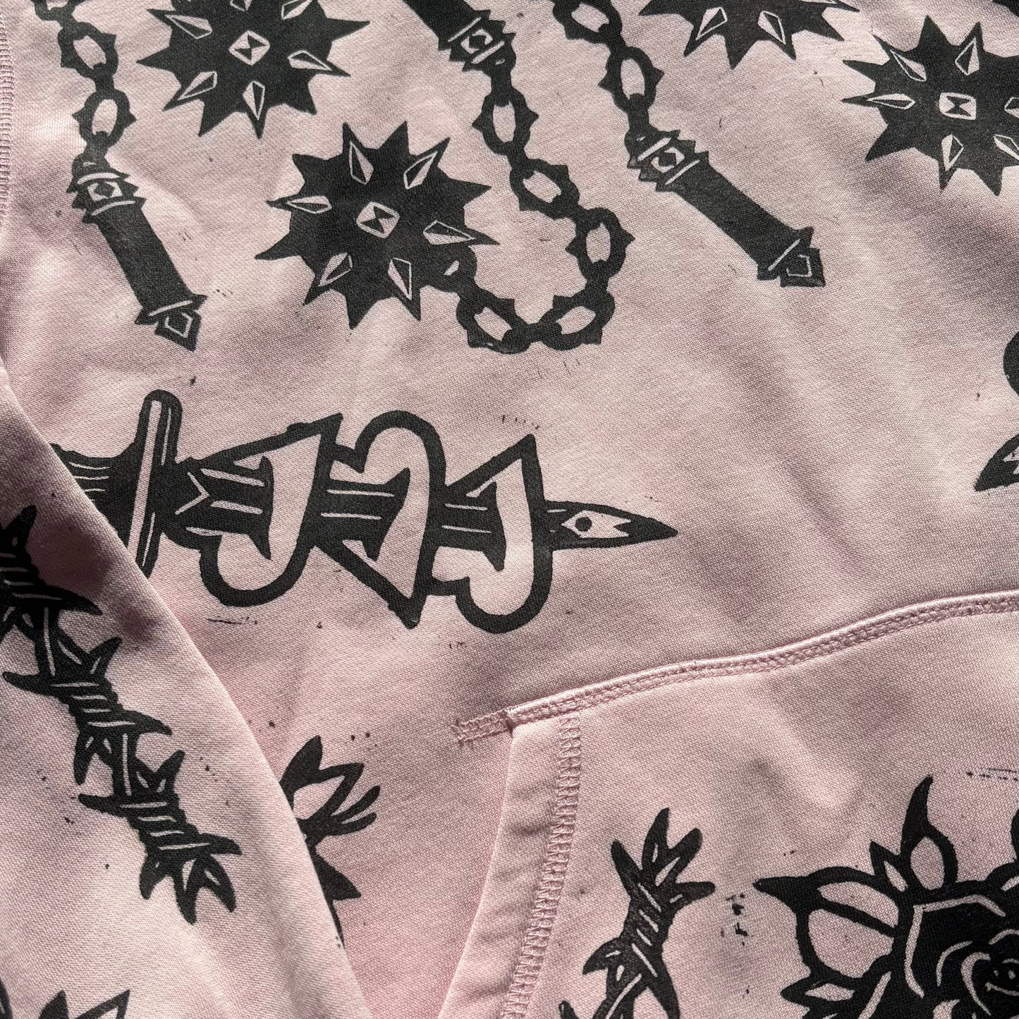 A close-up of a light pink hoodie featuring a black graphic print of a spiked mace, a rose, and barbed wire.