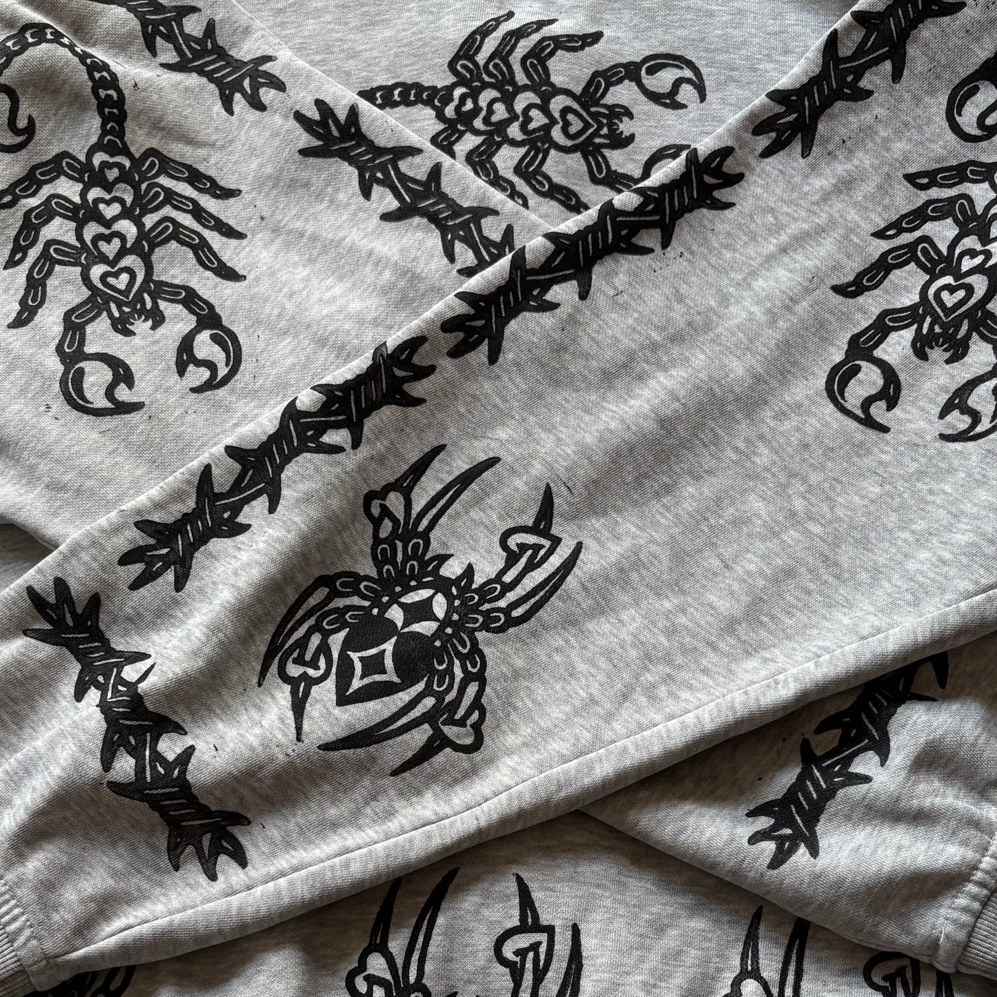 A close-up of a light gray heather sweatshirt featuring a black graphic print of a stylized scorpion and a black barbed wire pattern running along the sleeve seams.