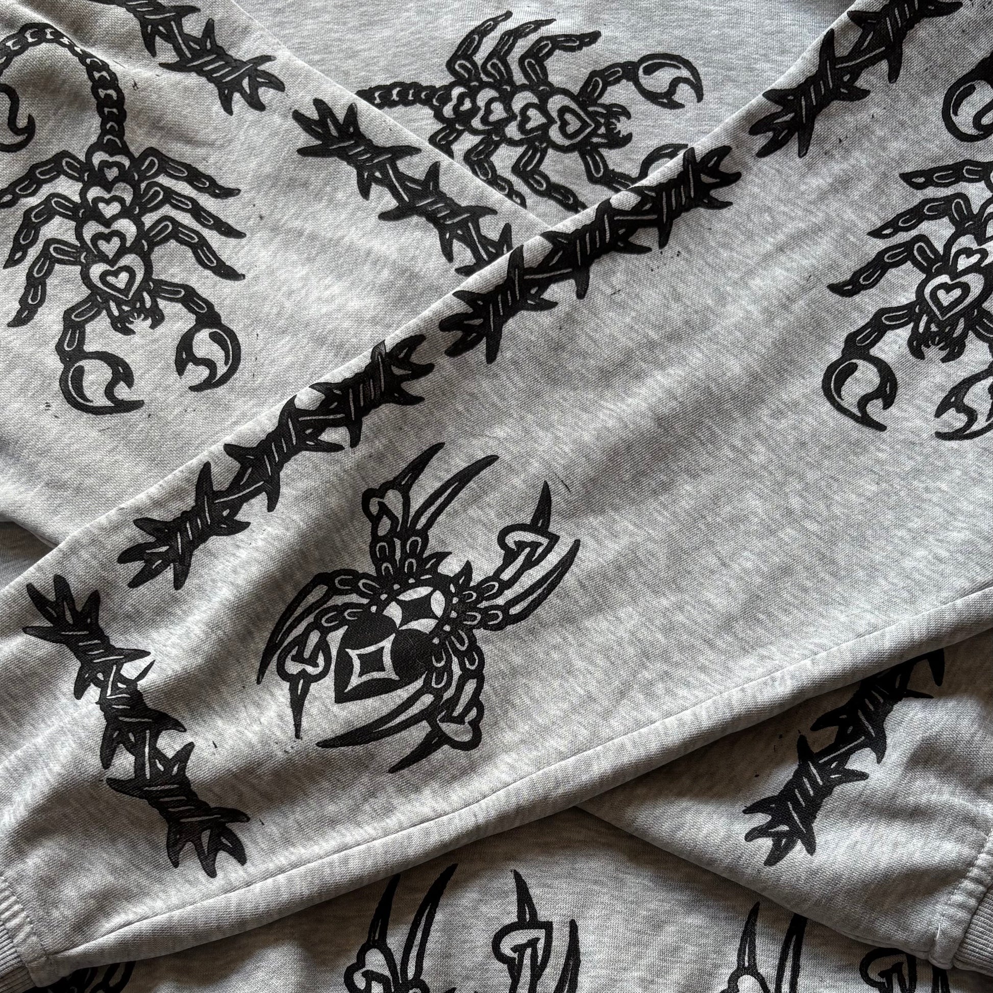 A close-up of a light gray heather sweatshirt featuring a black graphic print of a stylized scorpion and a black barbed wire pattern running along the sleeve seams.
