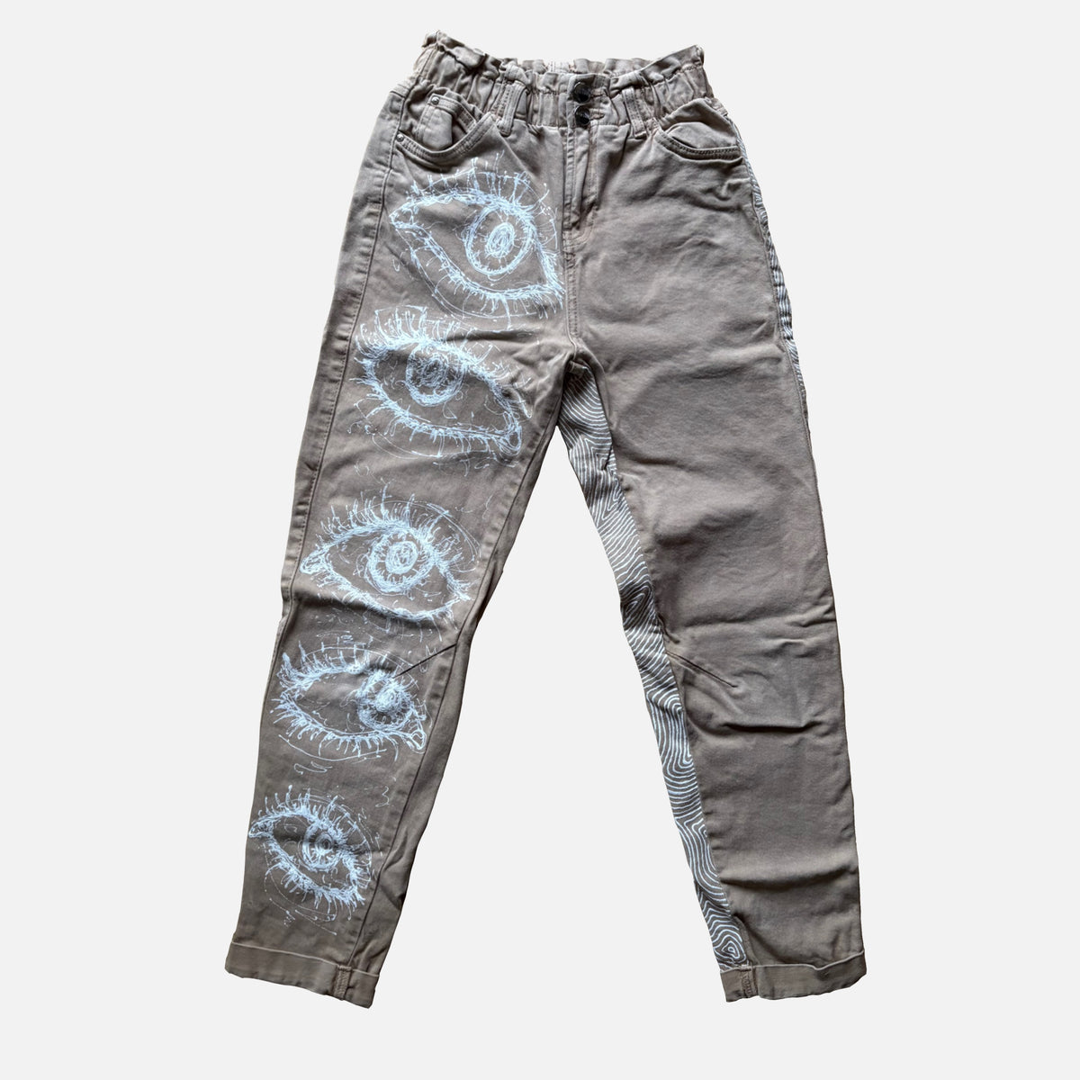 A pair of beige denim pants featuring a vertical series of white hand-drawn style eye illustrations on the left leg, with a matching eye pattern visible on the right leg's side seam.