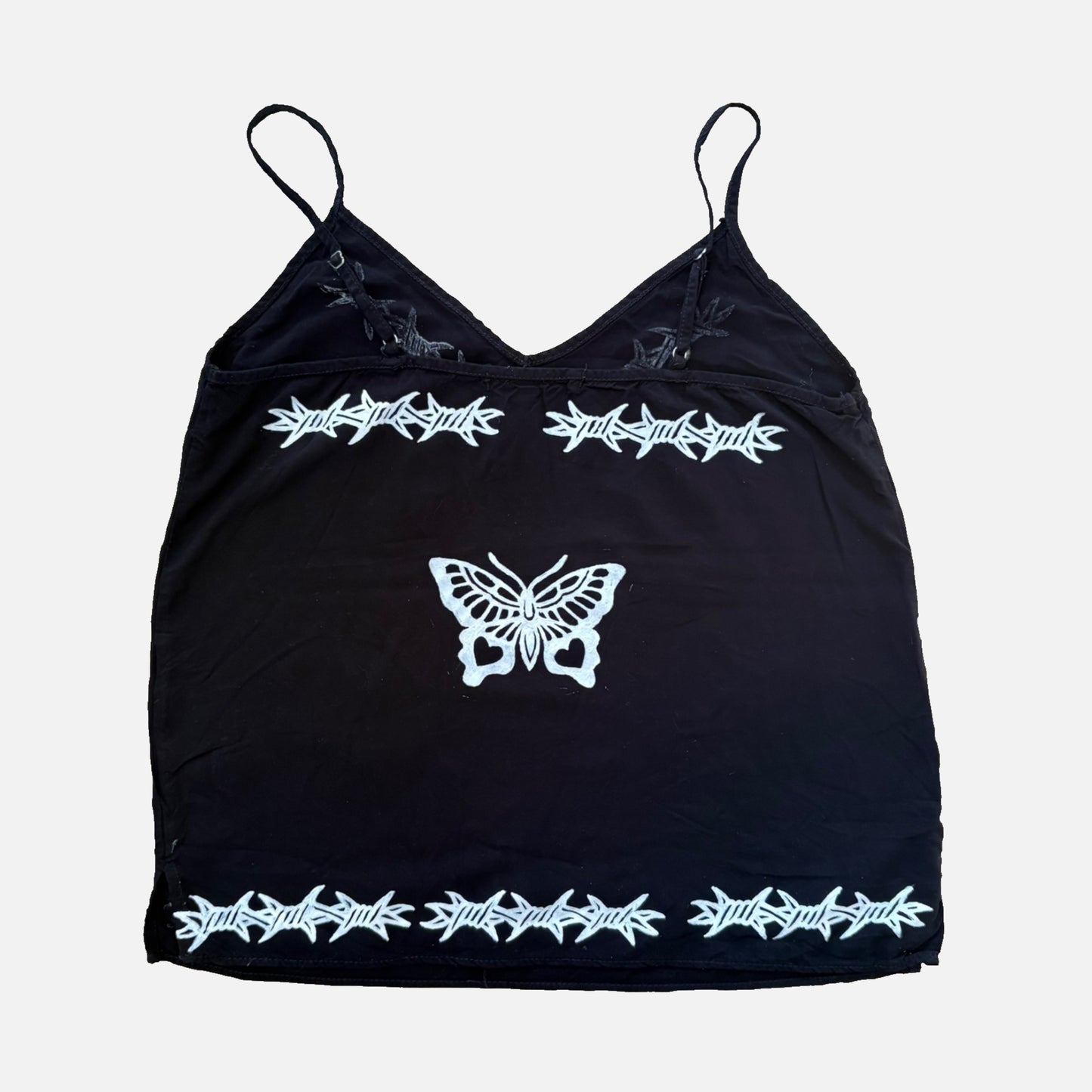 A black spaghetti-strap camisole top featuring white printed barbed wire designs across the chest, bottom hem, and center butterfly motif, shown against a light gray background.