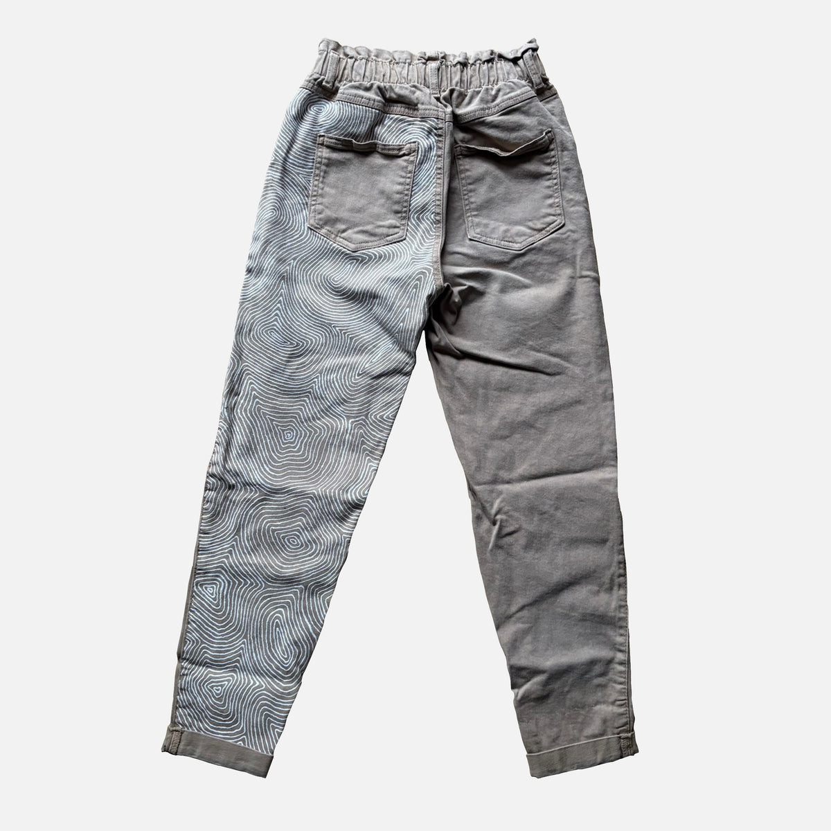 A pair of beige high-waisted pants with an elastic waistband, back pockets, and a wavy, organic pattern on the left leg, shown against a light gray background.