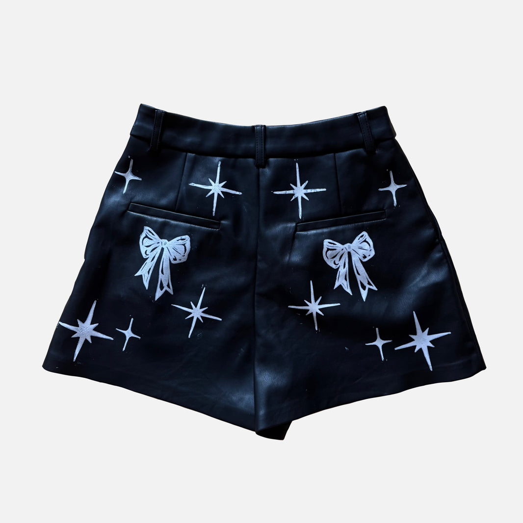 Black shorts with white printed starburst and bow patterns, shown flat against a light gray background.