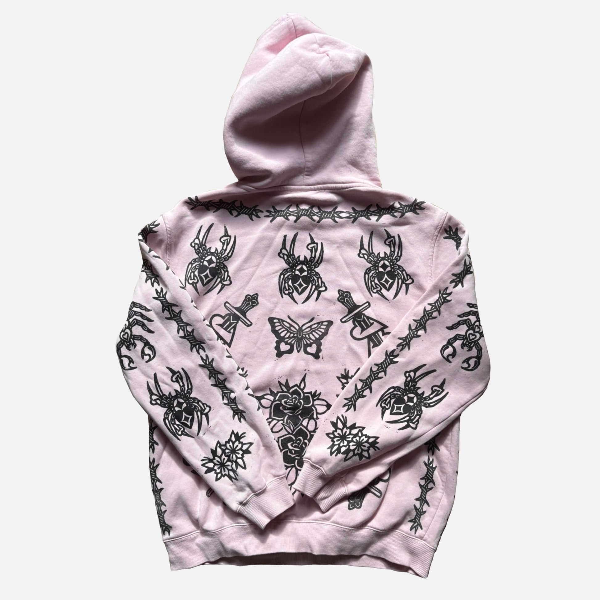 A light pink pullover hoodie with a black gothic graphic print featuring spiders, butterflies, roses, and barbed wire, shown against a plain light gray background.