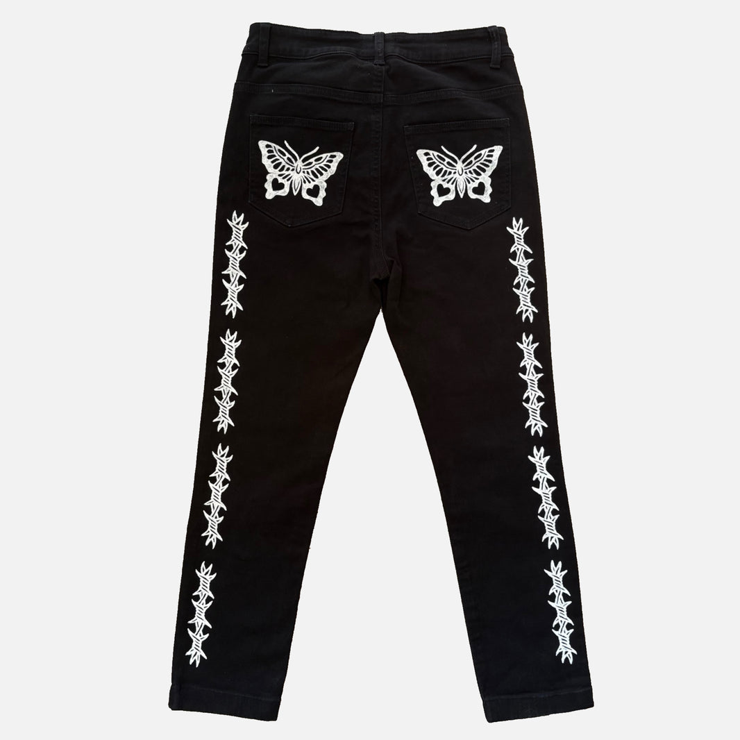 Black denim jeans with white printed butterfly designs on the back pockets and vertical white printed line patterns down both legs, shown against a light gray background.
