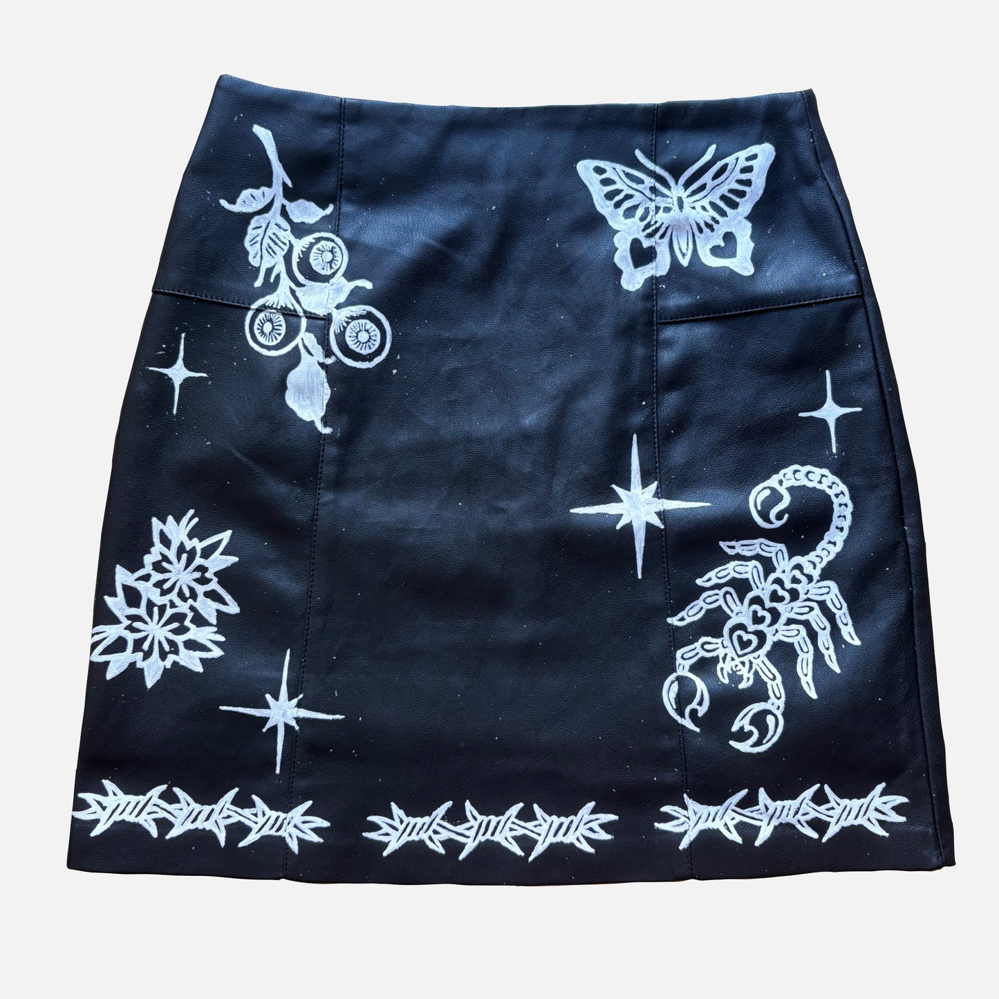 A black mini skirt featuring white gothic-style prints including a butterfly, scorpion, stars, a rose with thorns, and a barbed-wire border, shown flat against a light gray background.
