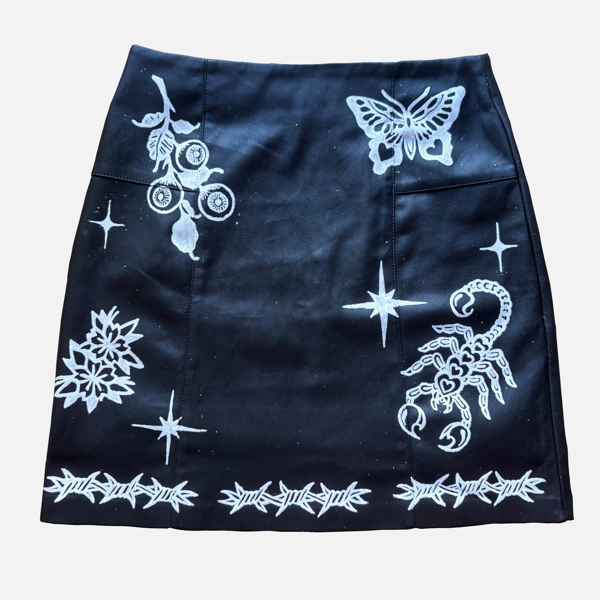 A black mini skirt featuring white gothic-style prints including a butterfly, scorpion, stars, a rose with thorns, and a barbed-wire border, shown flat against a light gray background.