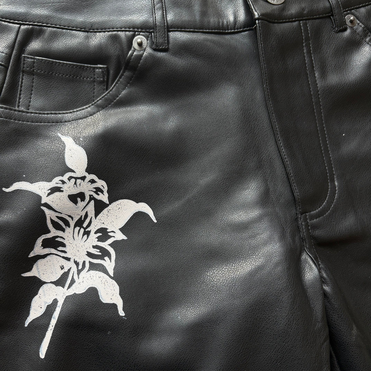 Close-up of black leather pants with a white graphic print of a flower and leaf on the left leg, showing the front pocket and metal rivets.