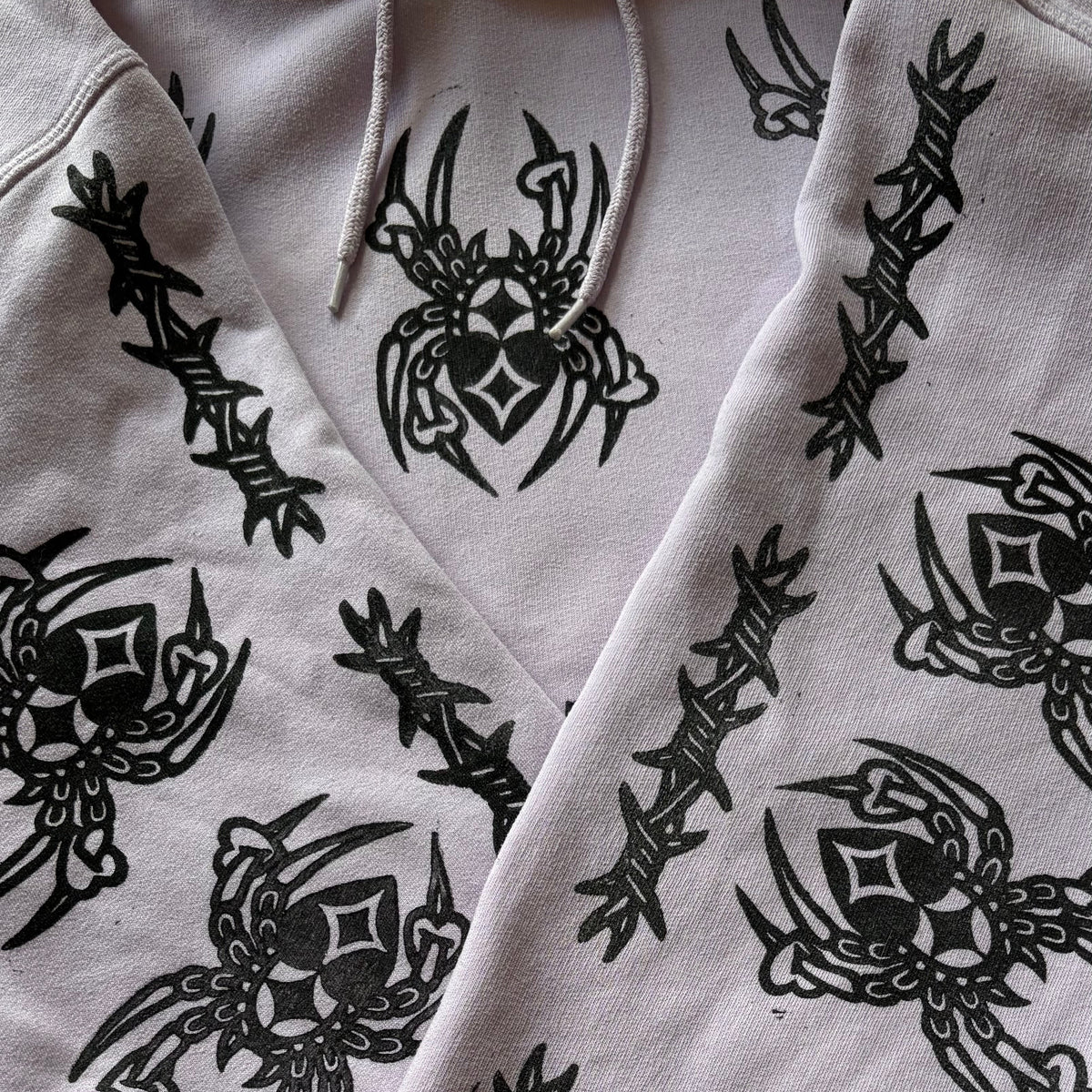 A close-up of a light purple hoodie featuring an all-over black graphic print of spiders and barbed wire.