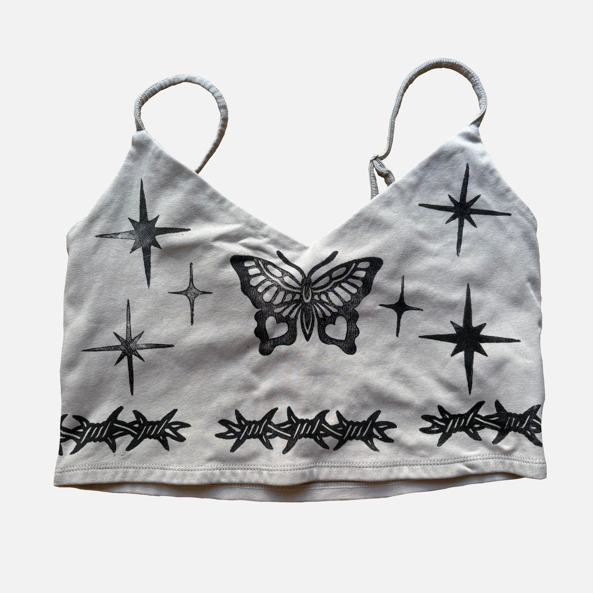 A beige cropped camisole top featuring black graphic prints of a butterfly, starbursts, and barbed wire, displayed against a light gray background.