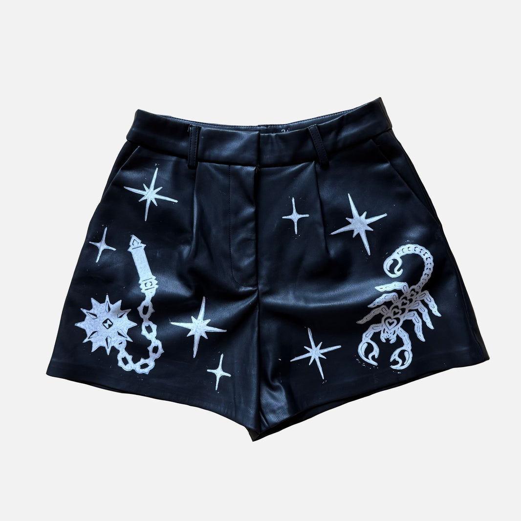 Black faux leather shorts featuring white printed celestial and astrological motifs, including stars, a scorpion, and a flail, shown against a light gray background.
