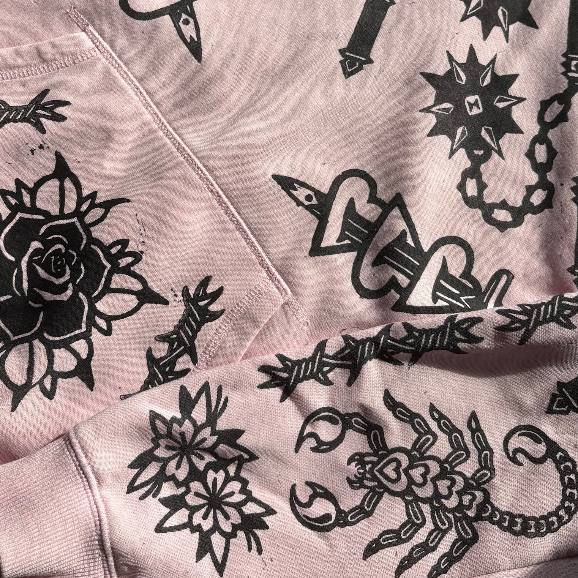 A close-up of a light pink hoodie featuring a black tattoo-style graphic print with roses, scorpions, chains, and knives.