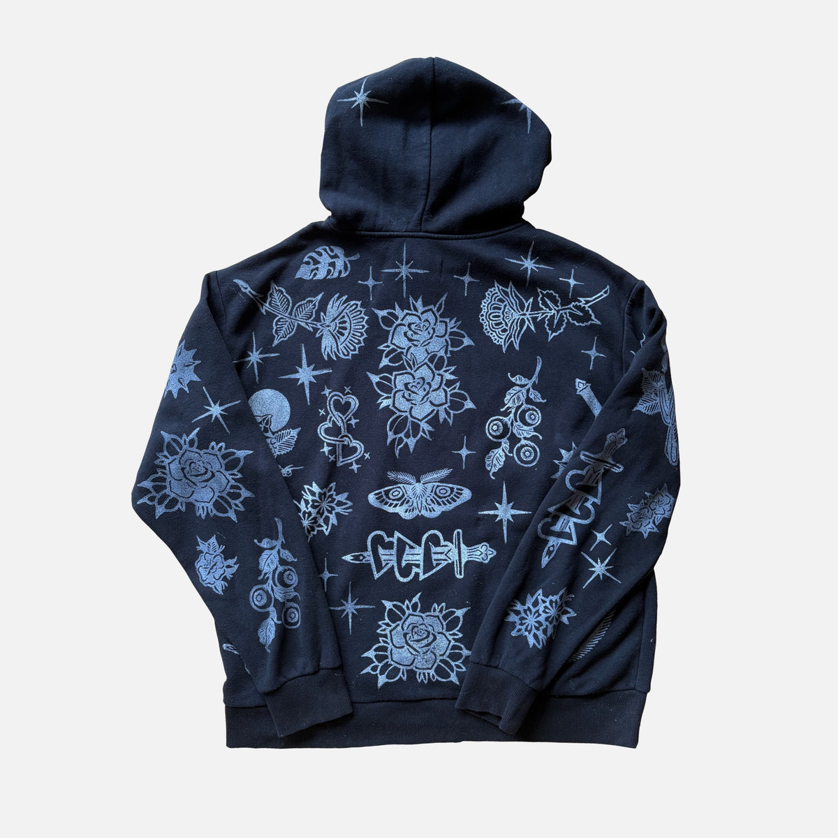A black hoodie shown from the back, featuring an all-over white graphic print of roses, stars, and occult-style symbols, displayed against a light gray background.