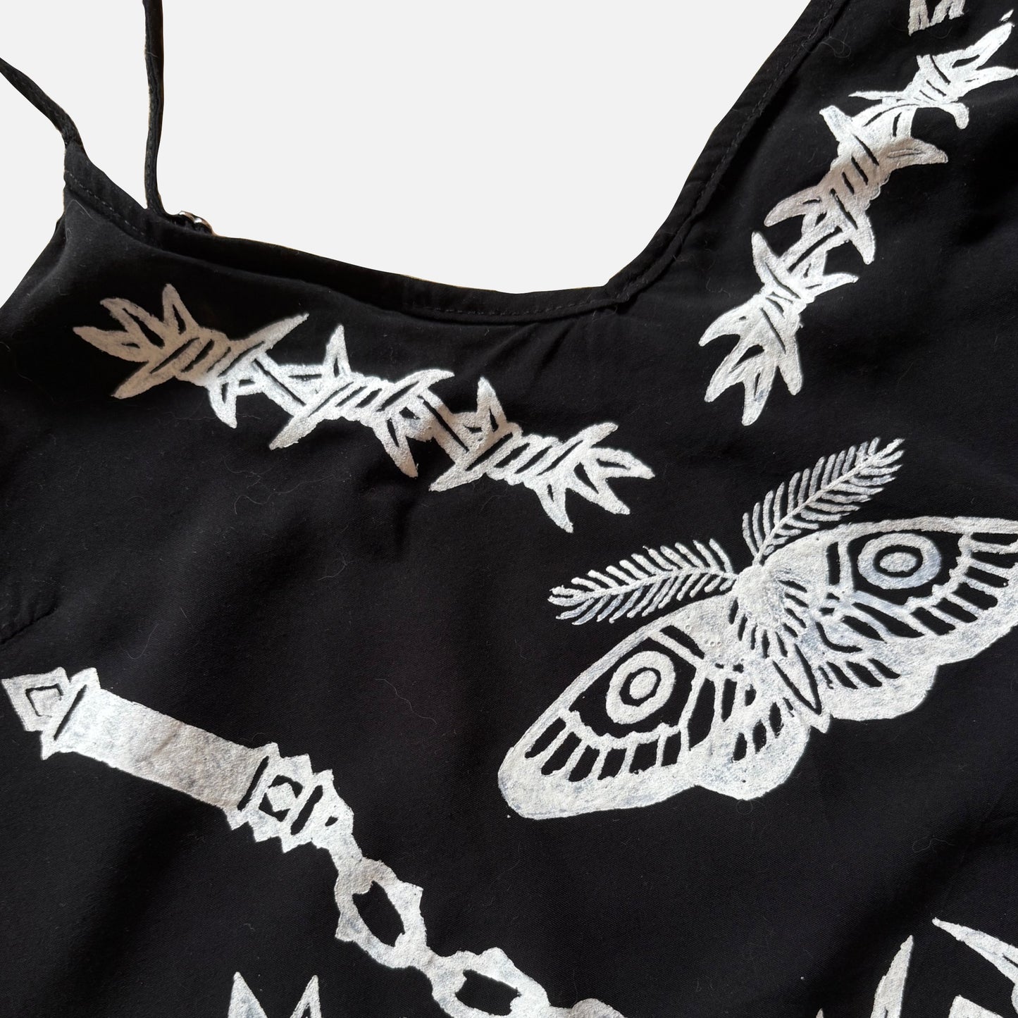 Close-up of a black garment featuring white printed barbed wire, a large moth motif, and a chain design on a light gray background.