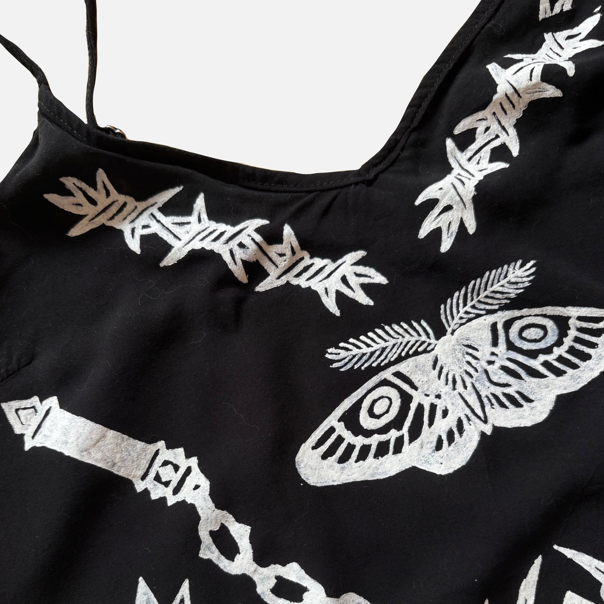 Close-up of a black garment featuring white printed barbed wire, a large moth motif, and a chain design on a light gray background.