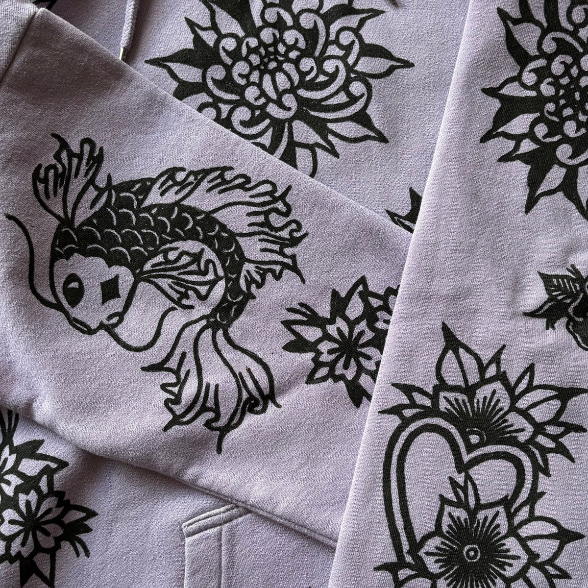 A close-up of a light purple hoodie featuring black line-art prints of a koi fish, lotus flowers, and a heart with floral details.