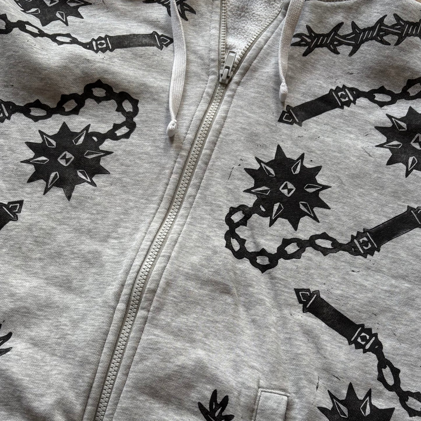 A light gray heather zip-up hoodie featuring a black graphic print of flails laid flat on a light surface.