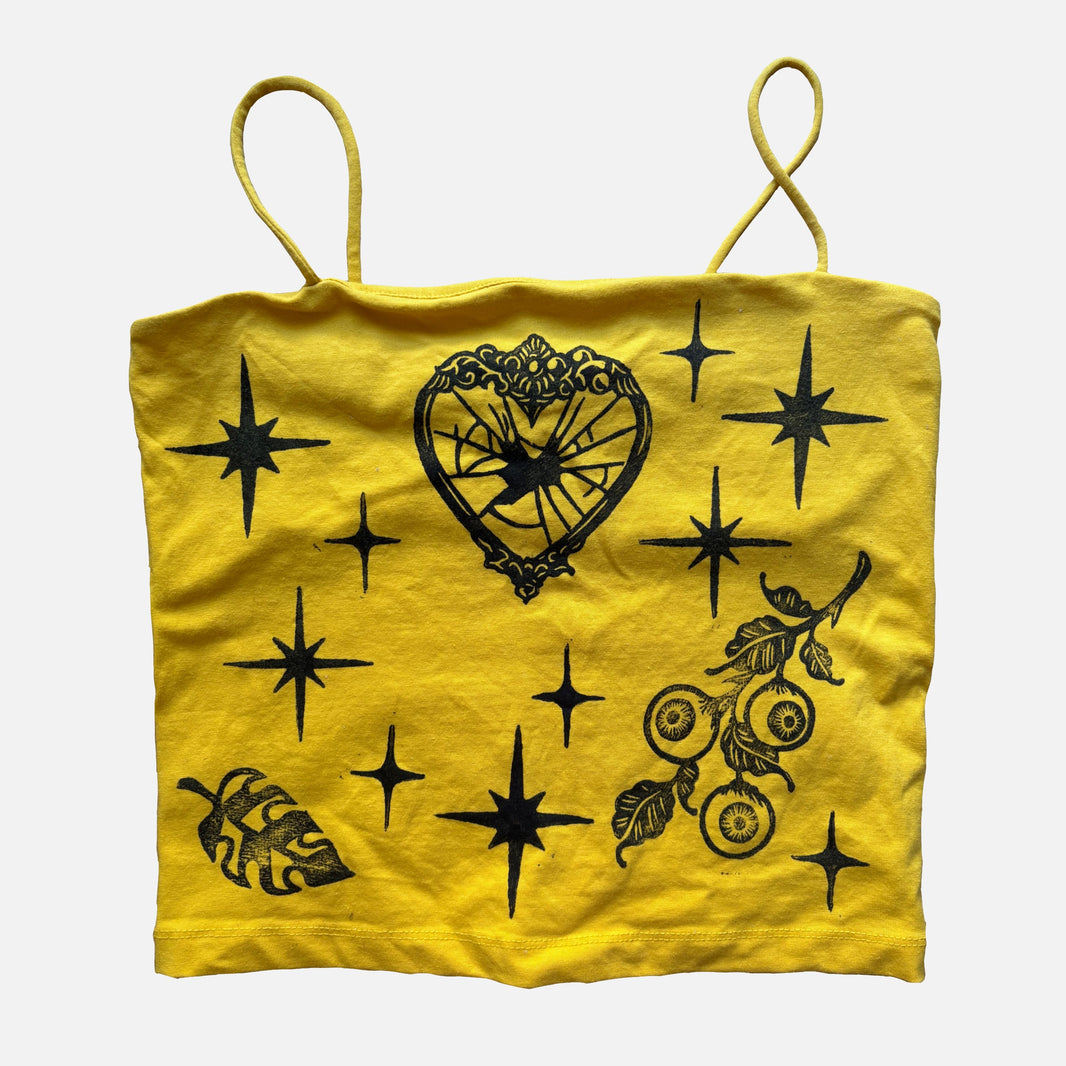 A yellow fabric crop top with spaghetti straps, featuring black printed graphics including stars, a heart-shaped mirror motif, a leaf, and a floral branch, shown against a light gray background.