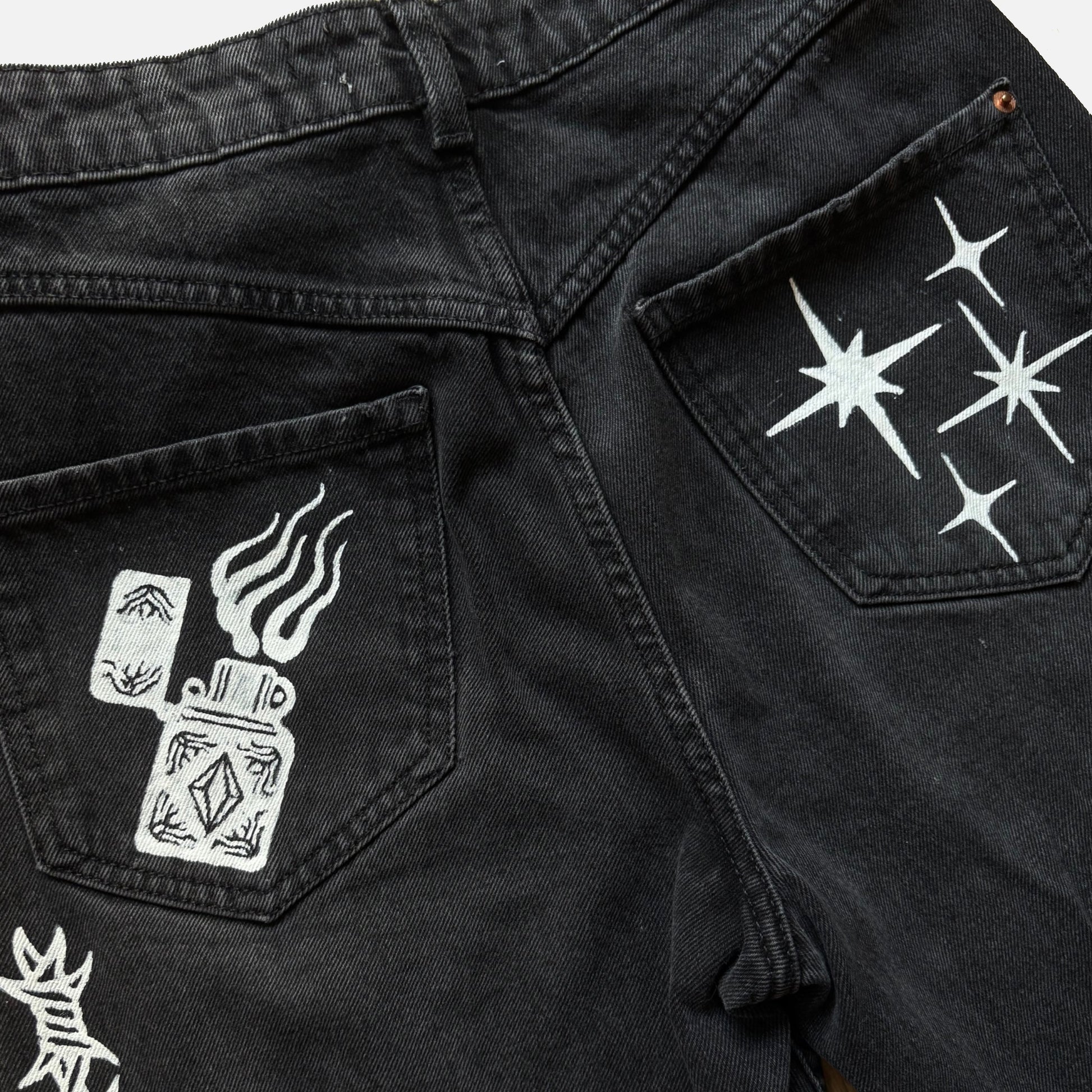 Close-up of black denim jeans showing the back pockets with white printed graphics, including a zippo lighter design on one pocket and starbursts on the other.