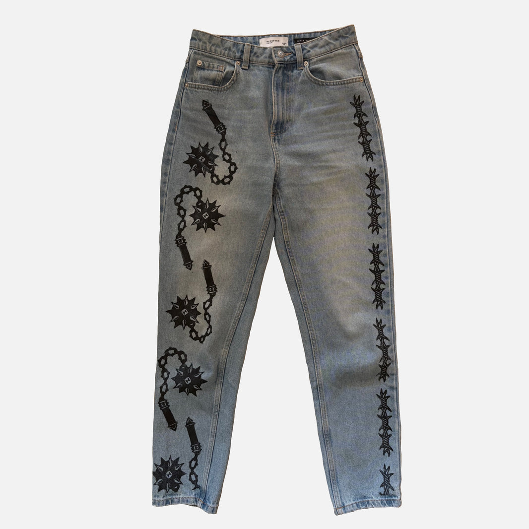 Light-wash blue denim jeans with black printed flail and barbed wire motifs running down both legs, shown flat against a light gray background.