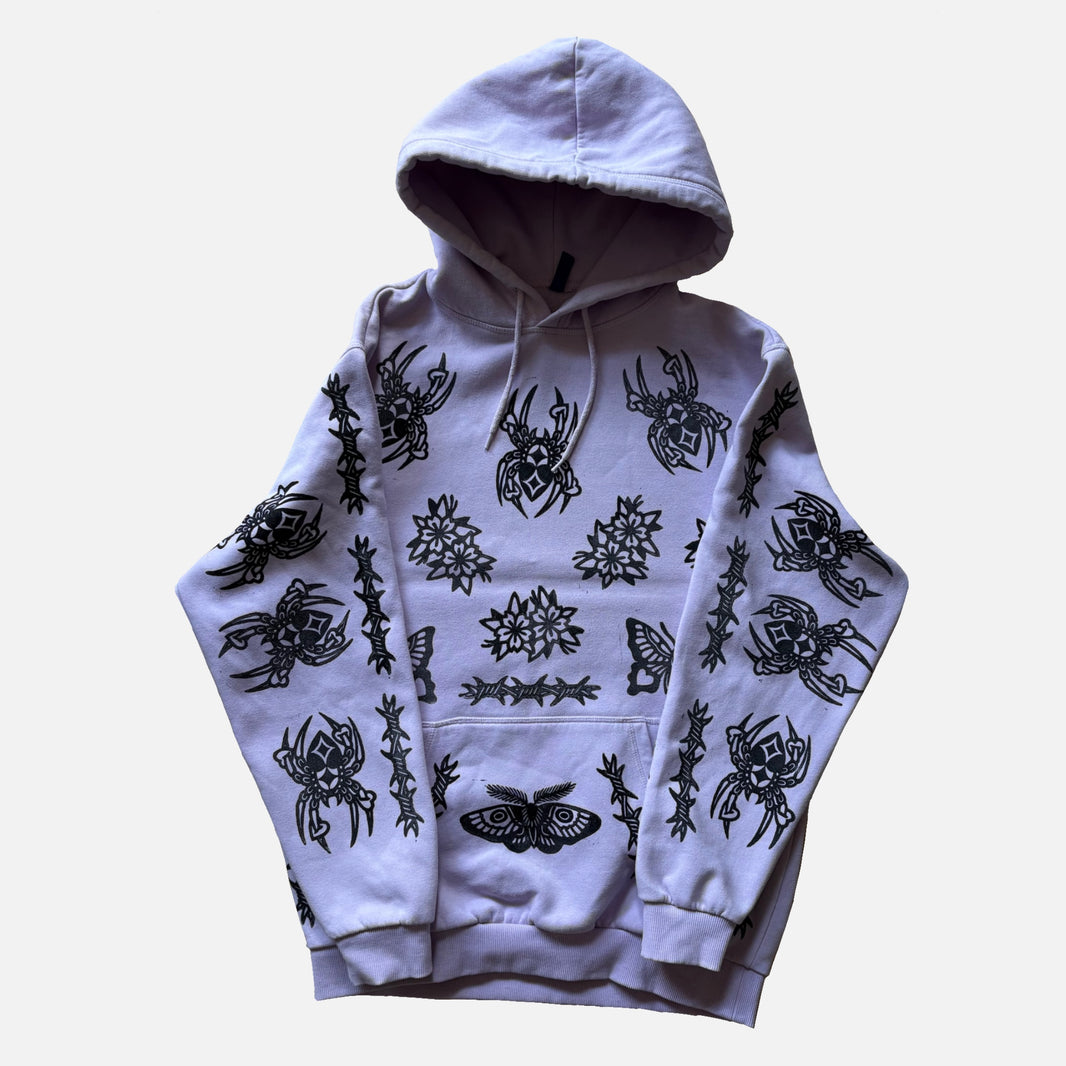 A light lavender-purple pullover hoodie featuring an all-over black graphic print of spiders, moths, flowers, and barbed wire designs, displayed against a plain light gray background.