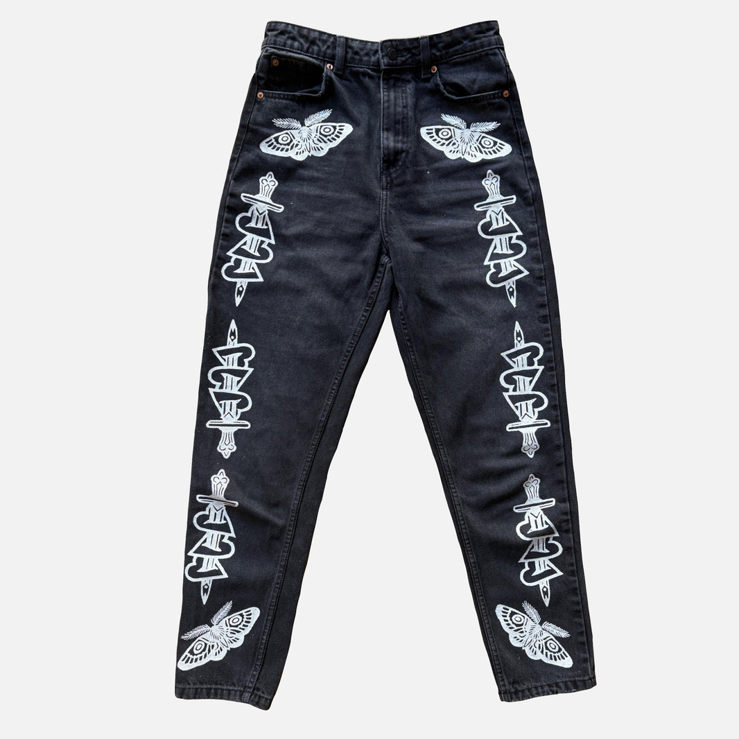 Black denim jeans featuring white printed motifs of moths and stacked knives down the outer thighs, displayed flat against a light gray background.