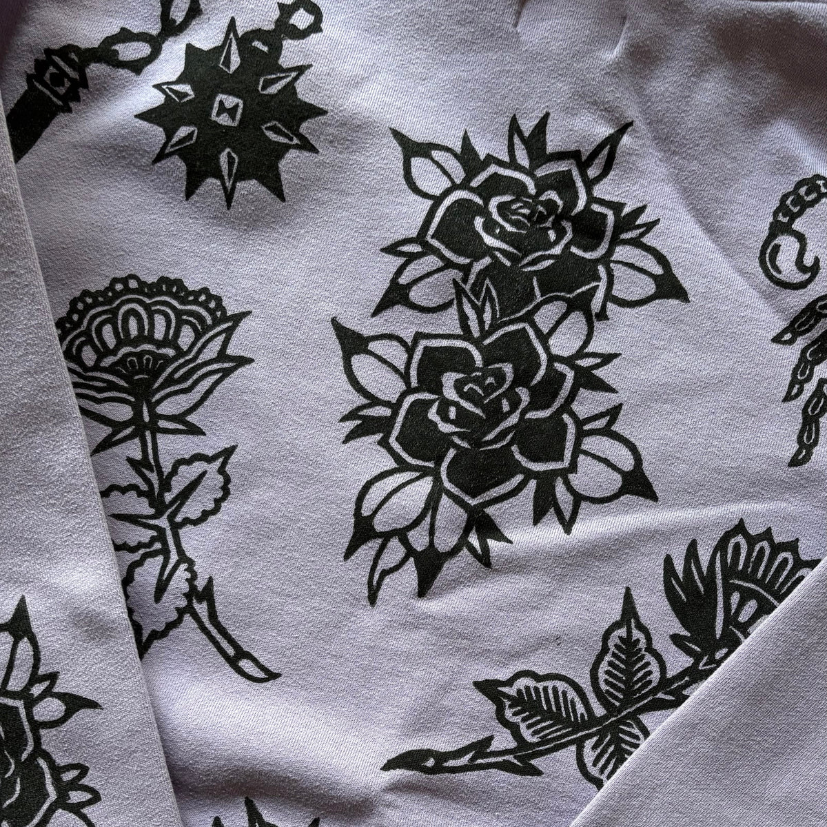 A close-up of a purple long-sleeve sweatshirt featuring black graphic prints of roses, flails, and a flowers, with ribbed cuffs and a crew neck.