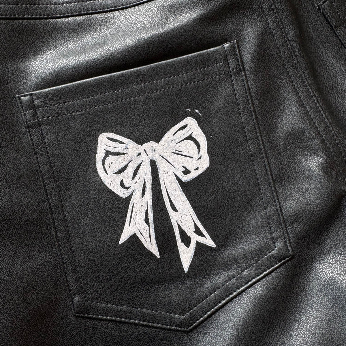 Close-up of black faux-leather pants showing the back pocket with a white hand-drawn bow graphic on it.