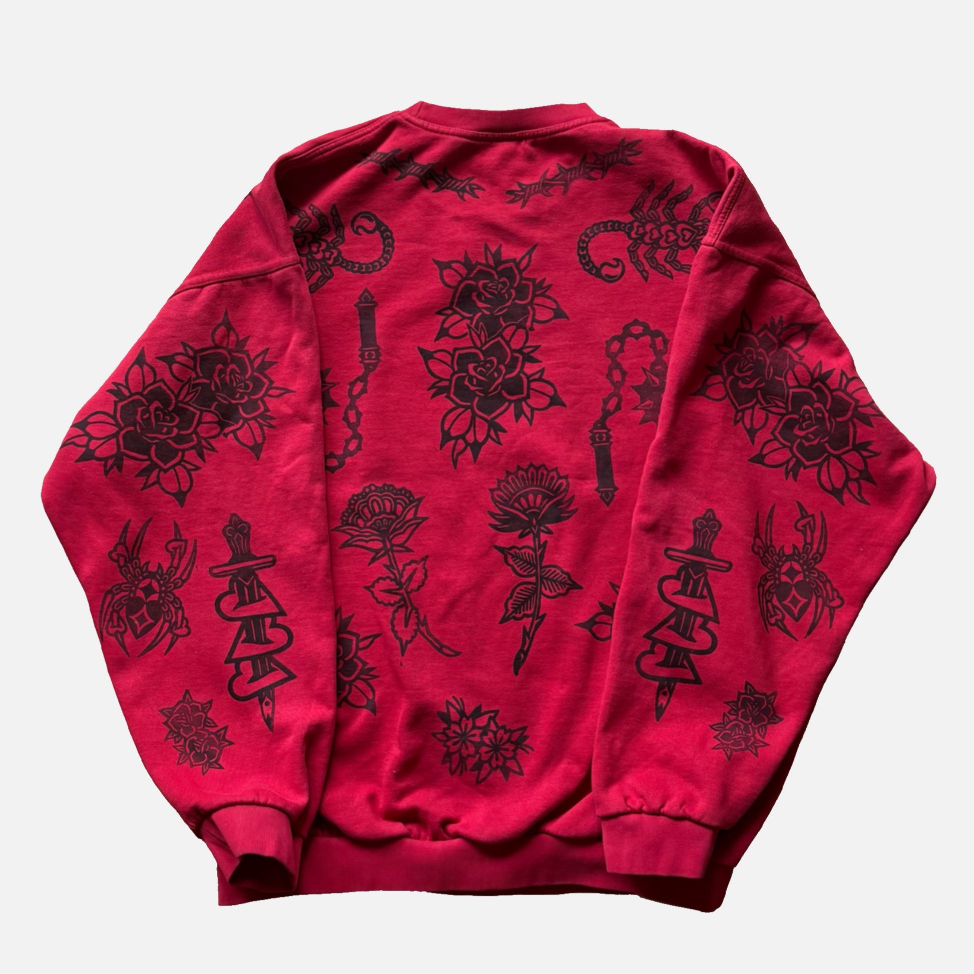 A red crewneck sweatshirt with black tattoo-style illustrations of roses, flowers, chains, and snakes, laid flat against a light gray background.