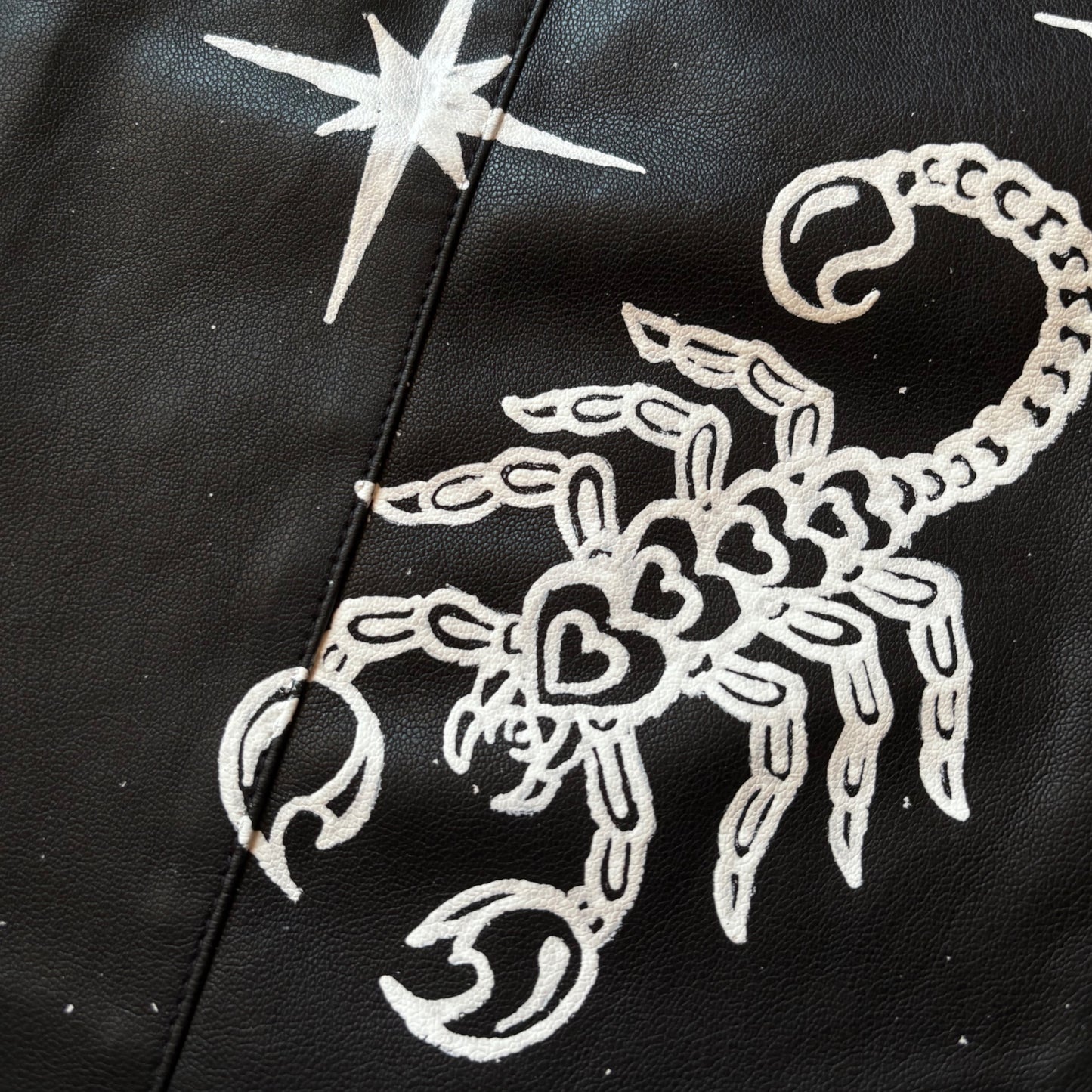 Close-up of a black leather surface featuring a white hand-painted scorpion design and a white star in the upper left.