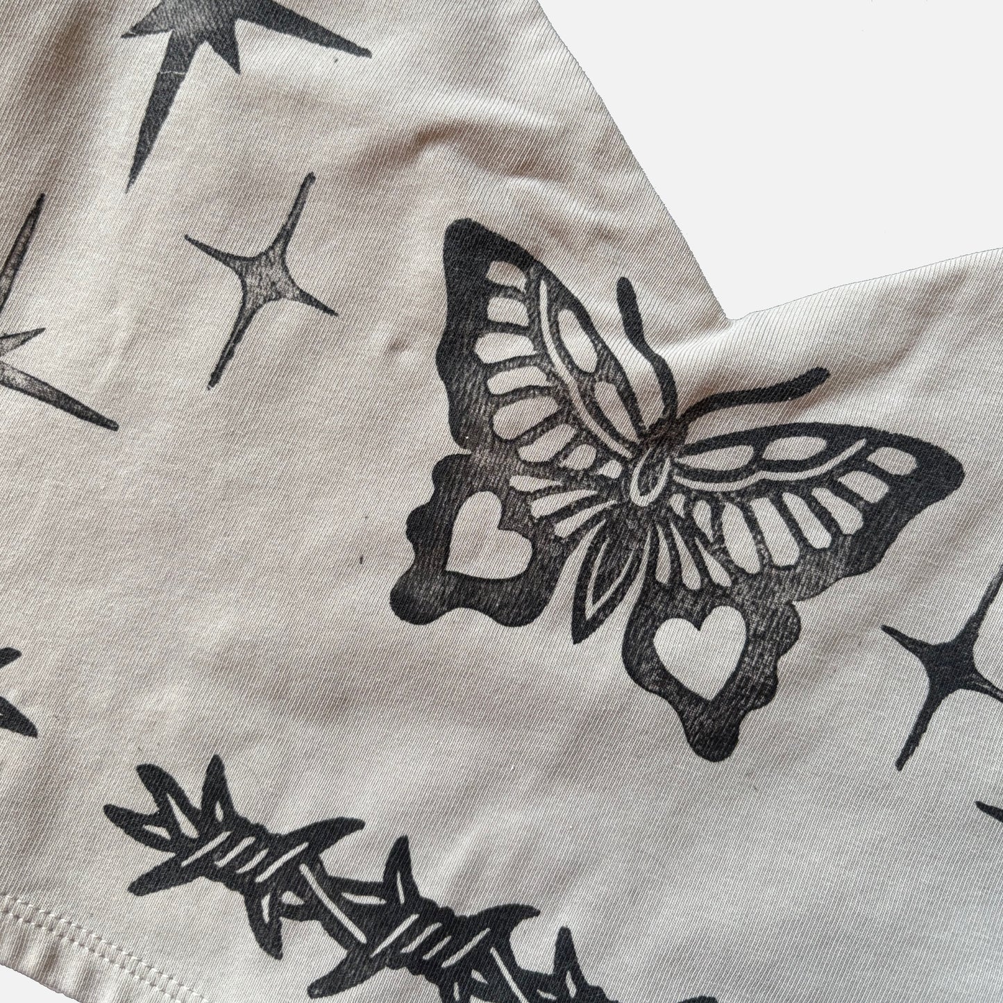 Close-up of a light beige fabric garment featuring black printed graphics, including a butterfly with heart-shaped cutouts, stars, and barbed wire.