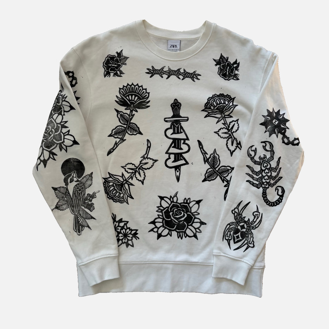 An off-white crewneck sweatshirt featuring black tattoo-style illustrations of roses, a dagger, a scorpion, a bird, and other graphic designs printed across the front and sleeves.