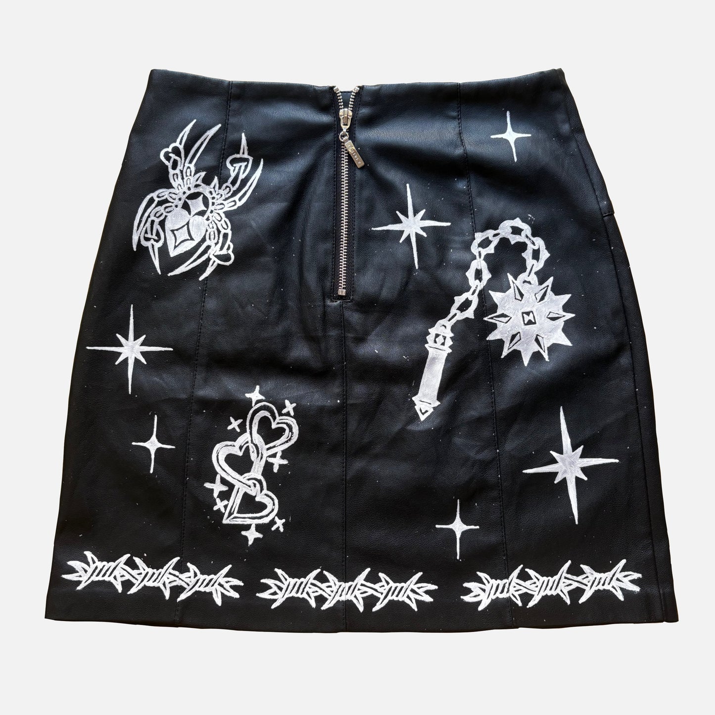 A black faux-leather mini skirt featuring white graphic prints of a flail, spider, a heart, and barbed wire, with a central zipper.