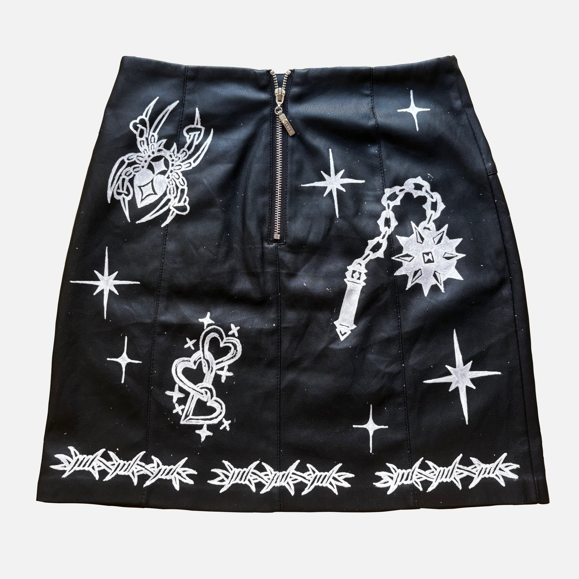 A black faux-leather mini skirt featuring white graphic prints of a flail, spider, a heart, and barbed wire, with a central zipper.