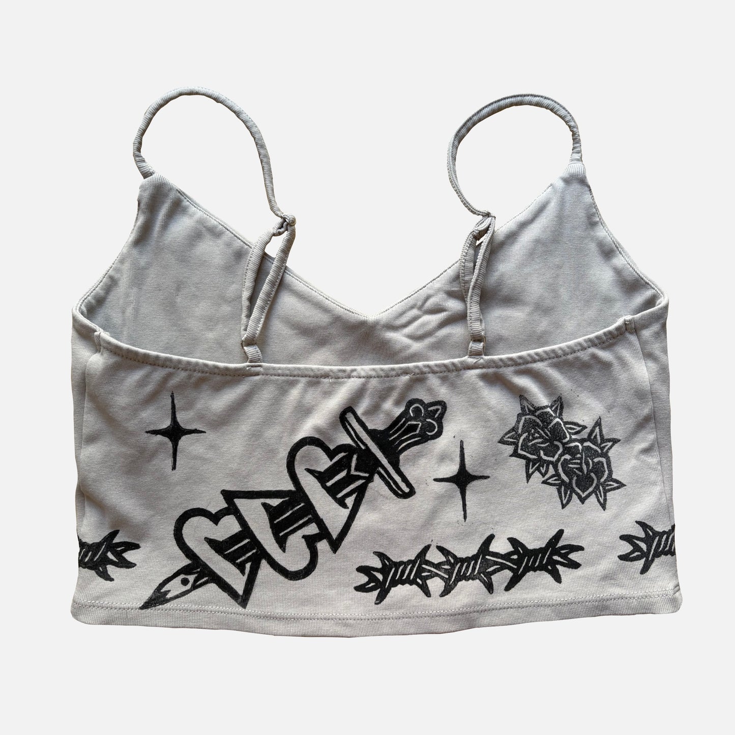 A beige cropped camisole top with black graphic prints of a dagger, roses, barbed wire, and stars, displayed against a light gray background.