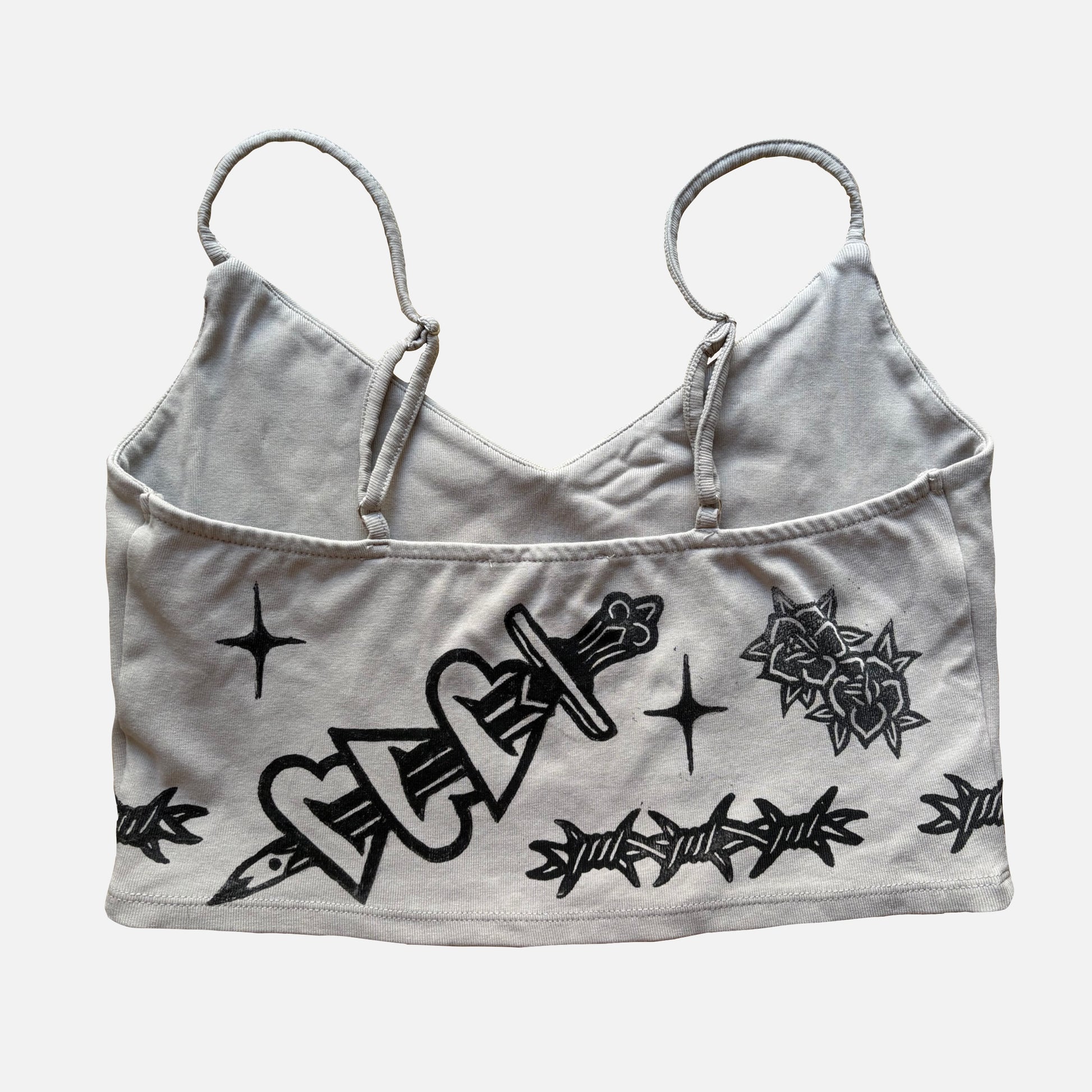 A beige cropped camisole top with black graphic prints of a dagger, roses, barbed wire, and stars, displayed against a light gray background.