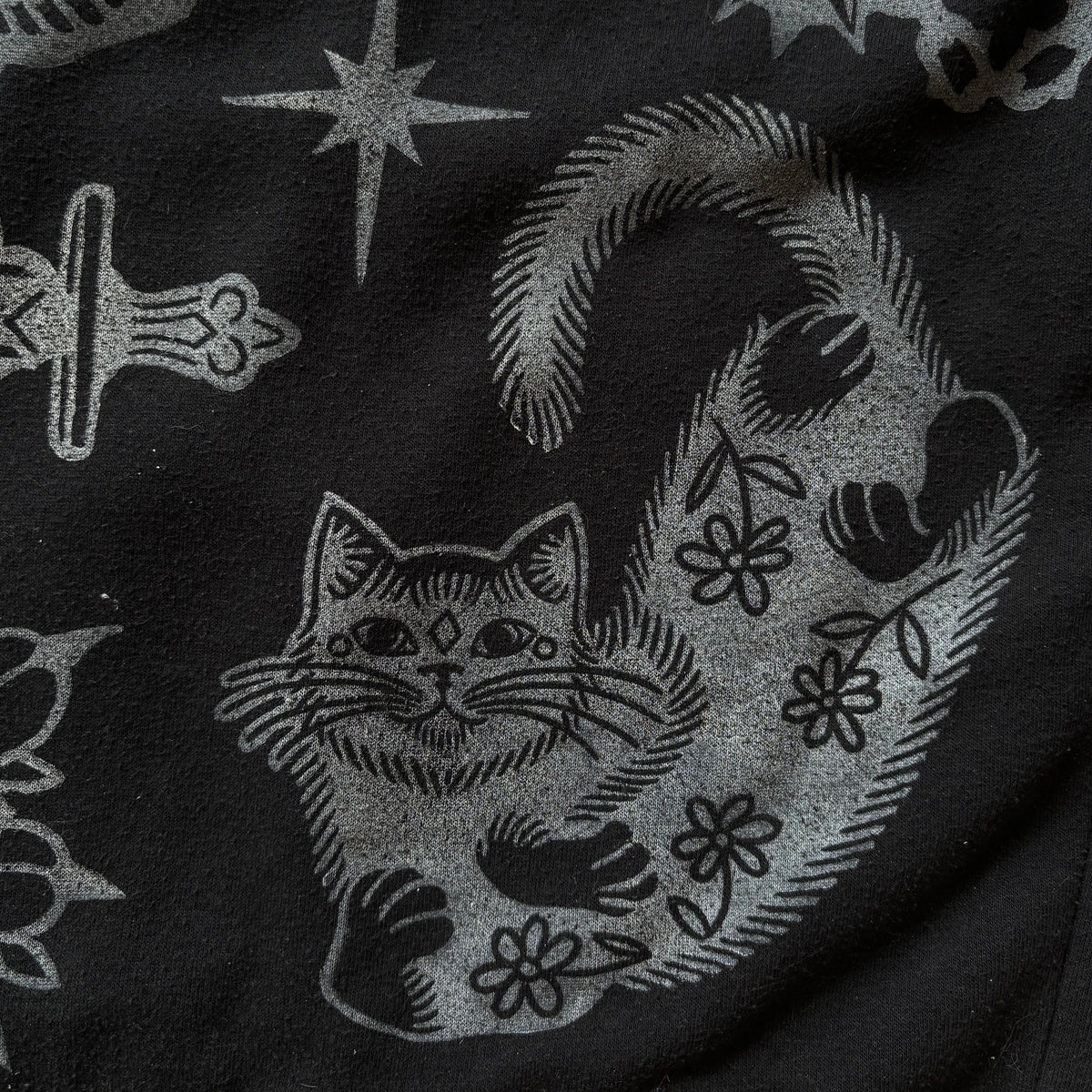 Close-up of a black fabric surface with a distressed white graphic print featuring a cat, flowers, stars, and occult-style symbols.