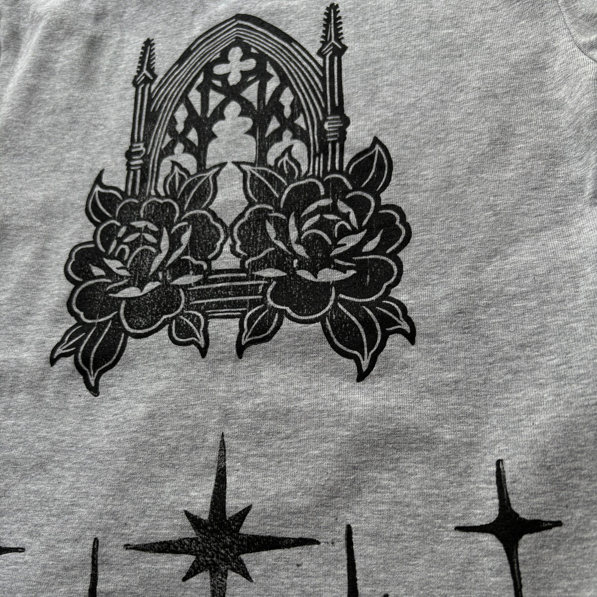 Close-up of a heather gray garment featuring a black graphic print of a gothic cathedral window framed by two large roses, with black starburst motifs below.
