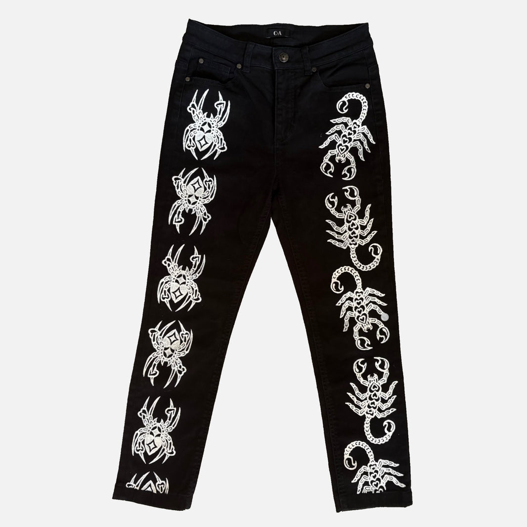 Black jeans with white printed spider and scorpion motifs running down both legs, shown flat against a light gray background.