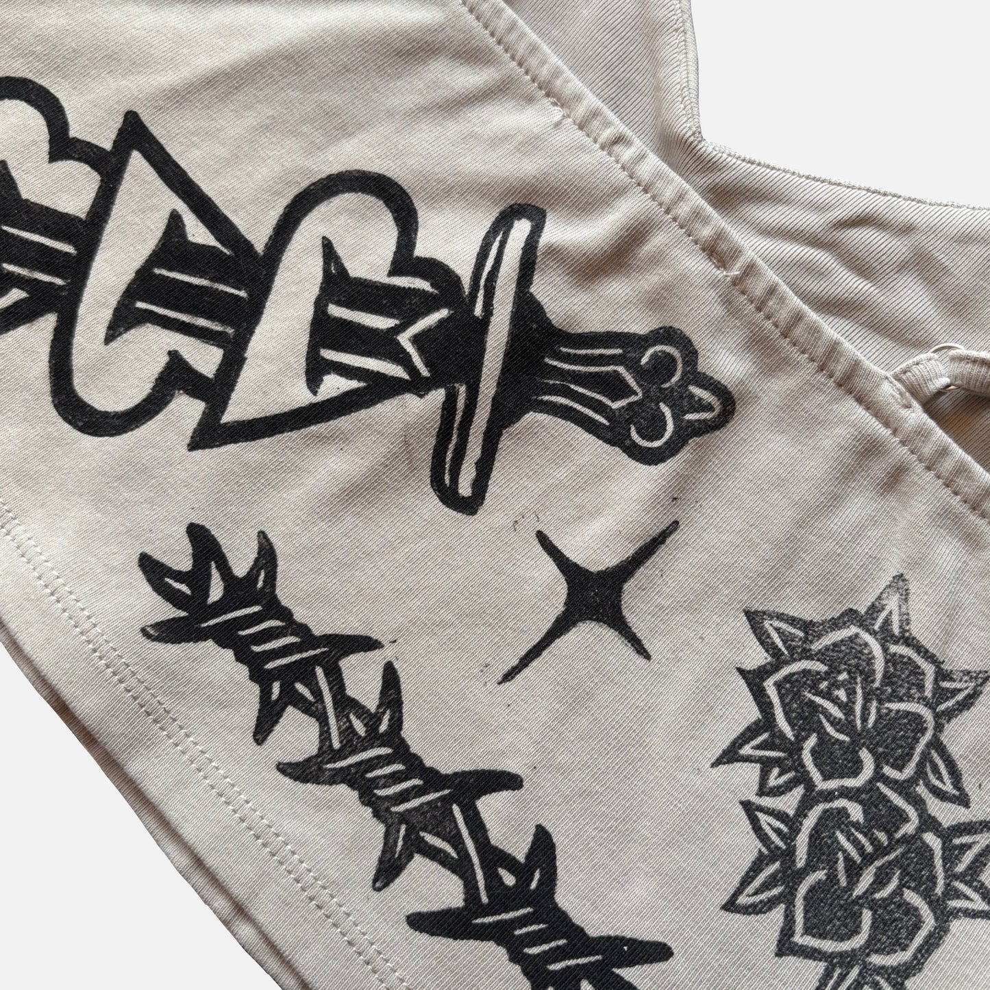 Close-up of a light beige fabric garment with black graphic prints, including a dagger, barbed wire, a star, and a rose.