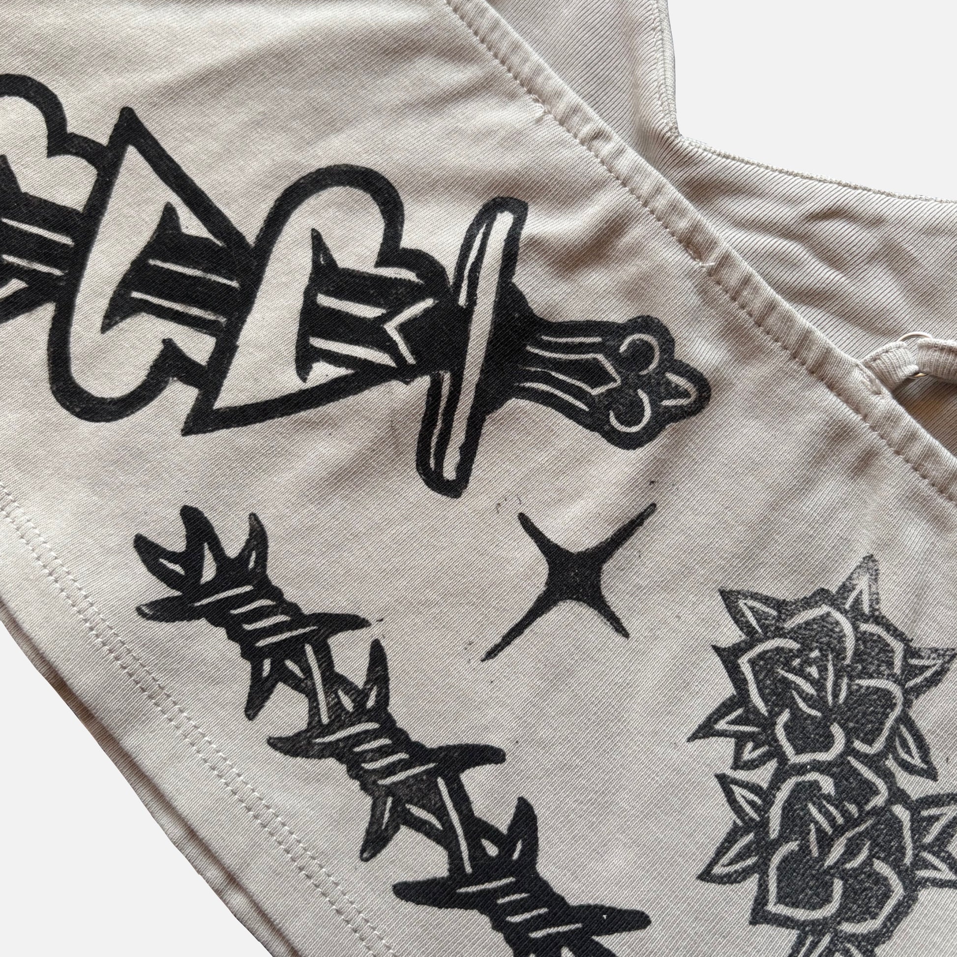 Close-up of a light beige fabric garment with black graphic prints, including a dagger, barbed wire, a star, and a rose.
