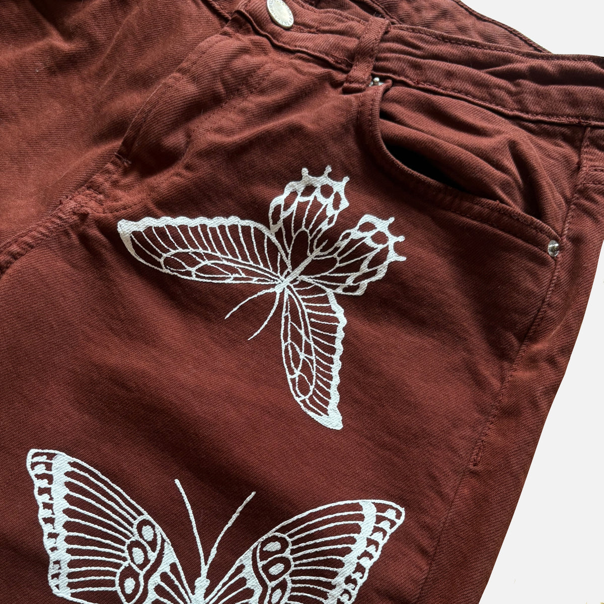 Close-up of a pair of rust-brown denim jeans featuring white painted butterfly designs on the front pocket.
